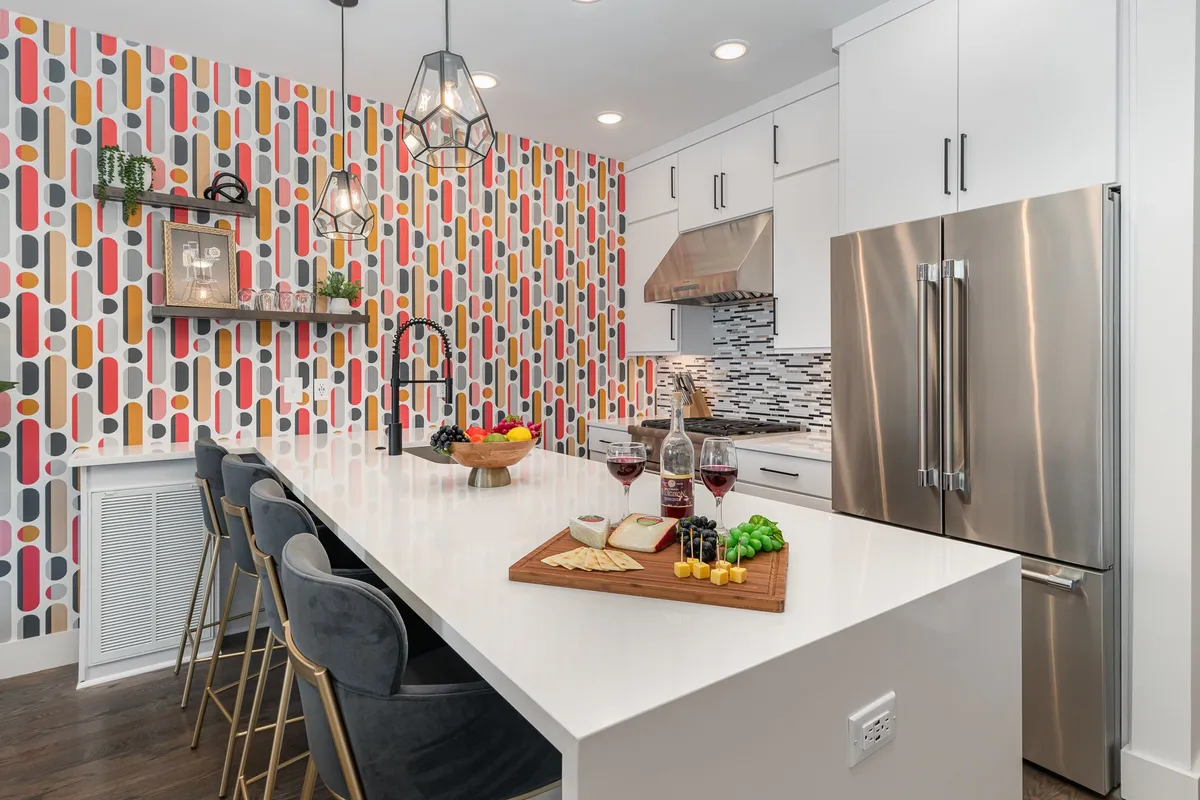 A cheerful, contemporary kitchen with colorful wallpaper as a backdrop, featuring a prominent kitchen island beautifully set with a food platter and wine glasses. The kitchen is equipped with modern appliances, and the island offers plenty of workspace and seating, suggesting both utility and sociability.