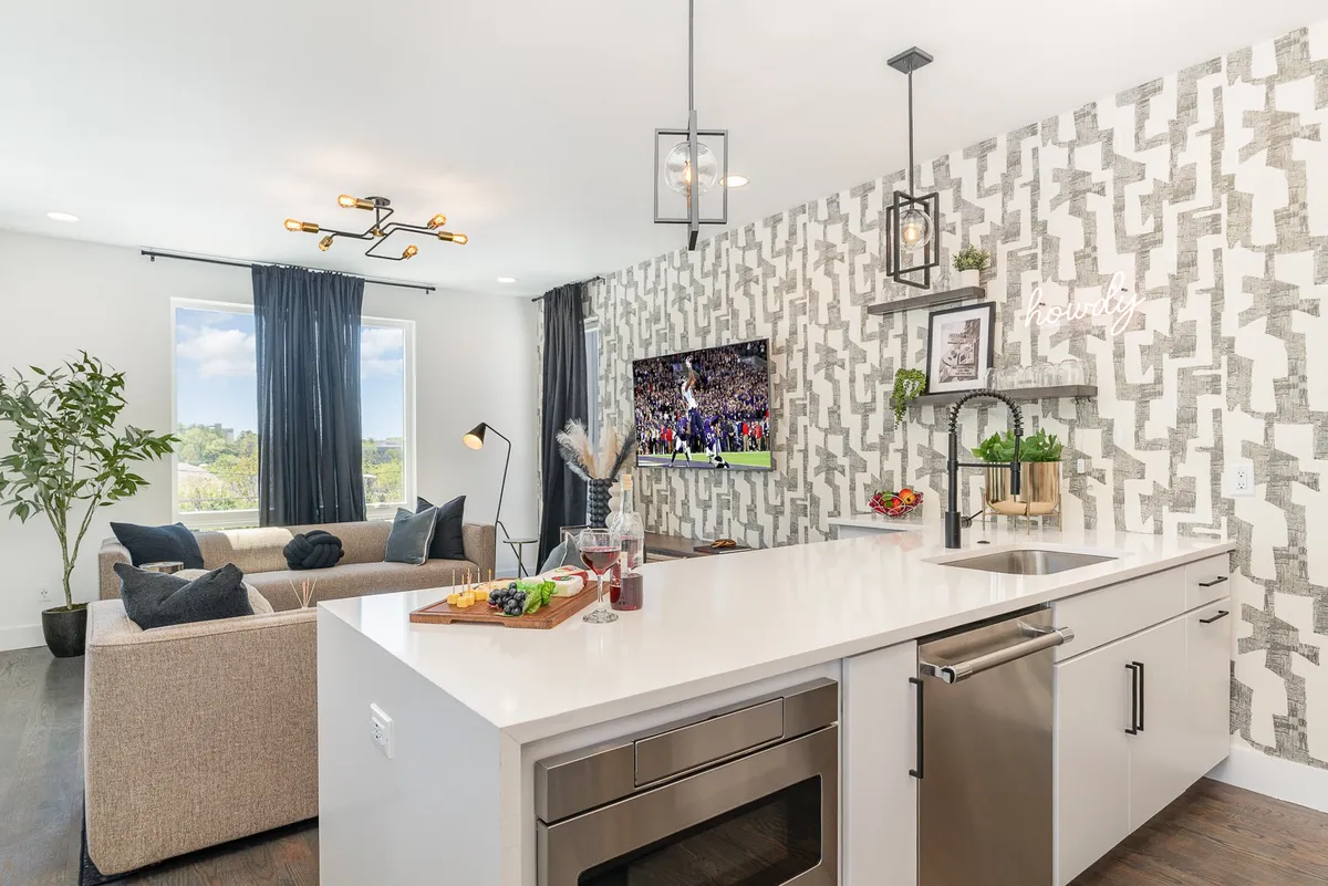 A spacious open-plan living area featuring a sleek kitchen island, plush sofa with decorative pillows, large window streaming with natural light, and a mounted TV showcasing a sports event. The room includes a decorative wall with geometric patterns, creating a lively and welcoming space.