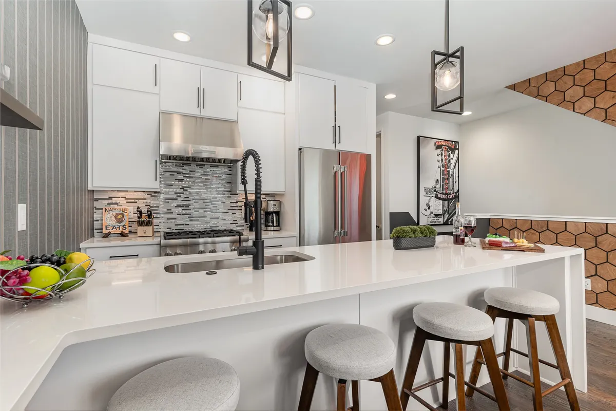 A welcoming, modern kitchen scene with an expansive island countertop, sleek bar stools, visible kitchen appliances, and artistic decorative elements. The space is illuminated by stylish light fixtures, and the setup encourages both cooking and comfortable group gatherings.