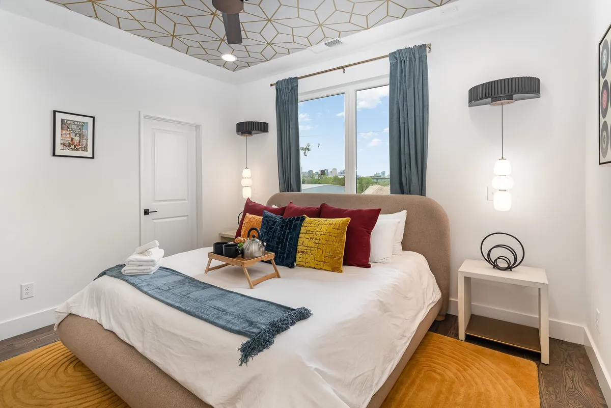 This image features a serene bedroom with a bed adorned with decorative pillows, a window with soft curtains allowing natural light to fill the space, tasteful wall art, and nightstands topped with lamps. The scene radiates calm and comfort, inviting relaxation or a quiet morning coffee.