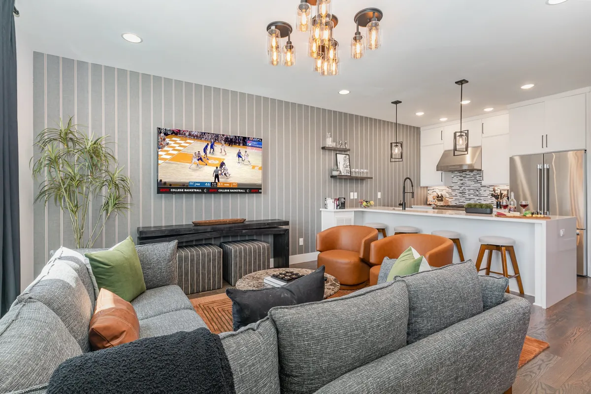 A welcoming, open-concept living area featuring a plush sectional sofa arranged around a modern coffee table, with a flat-screen TV mounted across from the seating. The kitchen island is visible in the background, seamlessly connecting the living and dining spaces. The room is bathed in warm light, creating a lively, inviting atmosphere ideal for group gatherings, watching movies, or celebrating special occasions.
