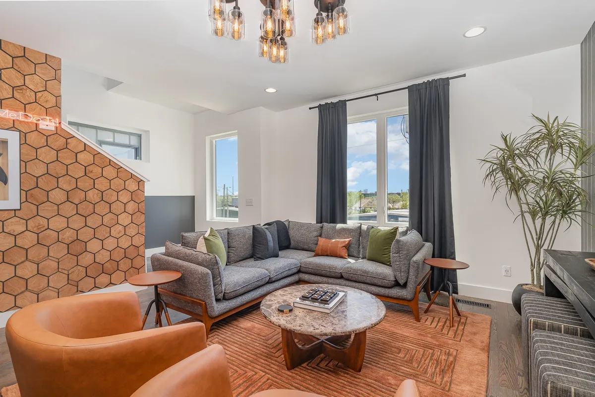 A cozy, welcoming living area with a large sectional sofa facing a chic coffee table. The hexagonal accent wall and stylish light fixture add character, while natural light streams through large windows, illuminating the space. The arrangement is ideal for shared moments, conversations, and leisurely afternoons.