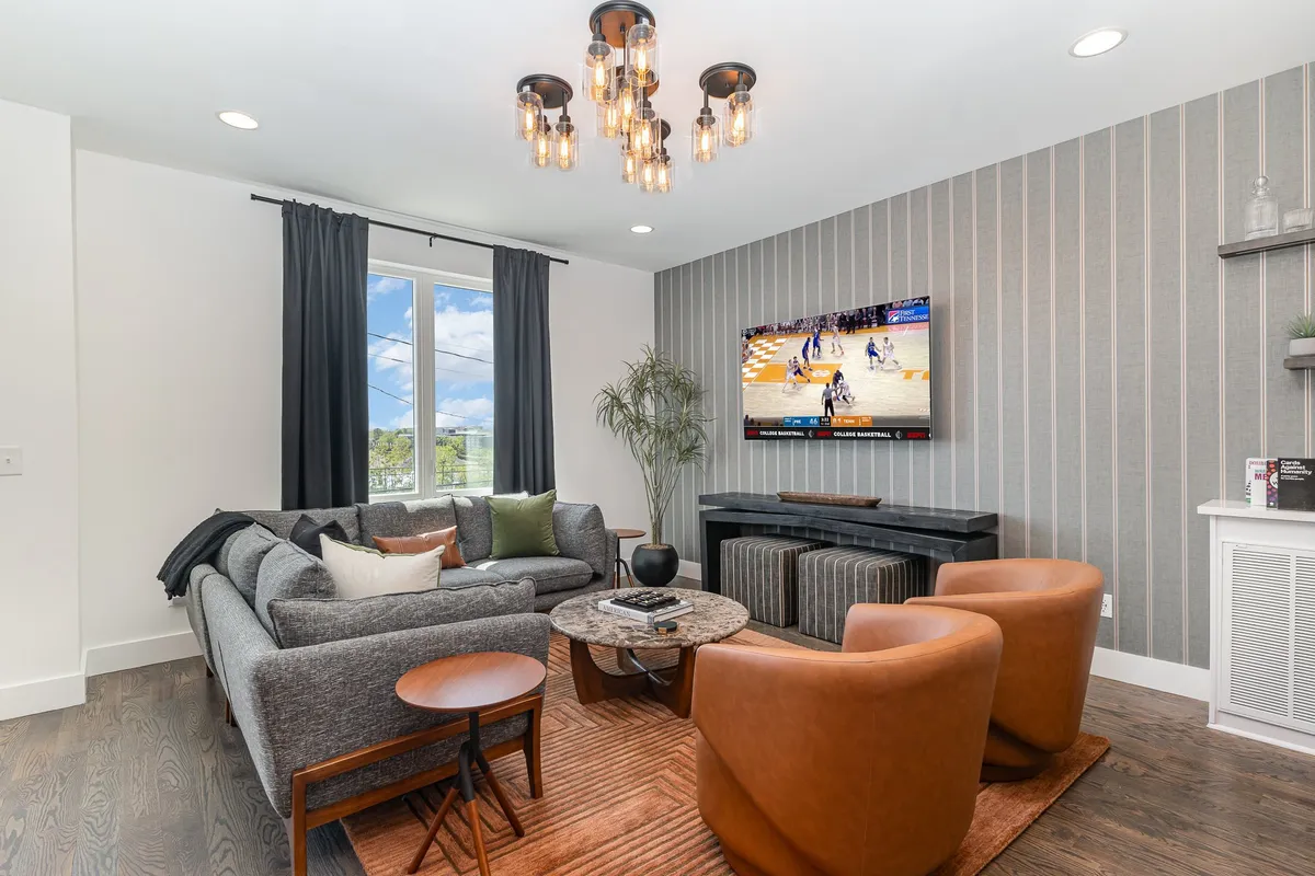 A modern, comfortable lounge space with a central sofa and armchairs, a TV displaying a sports game, a striking chandelier, and a decorative plant adding a touch of greenery. The atmosphere is inviting and encourages social interaction.