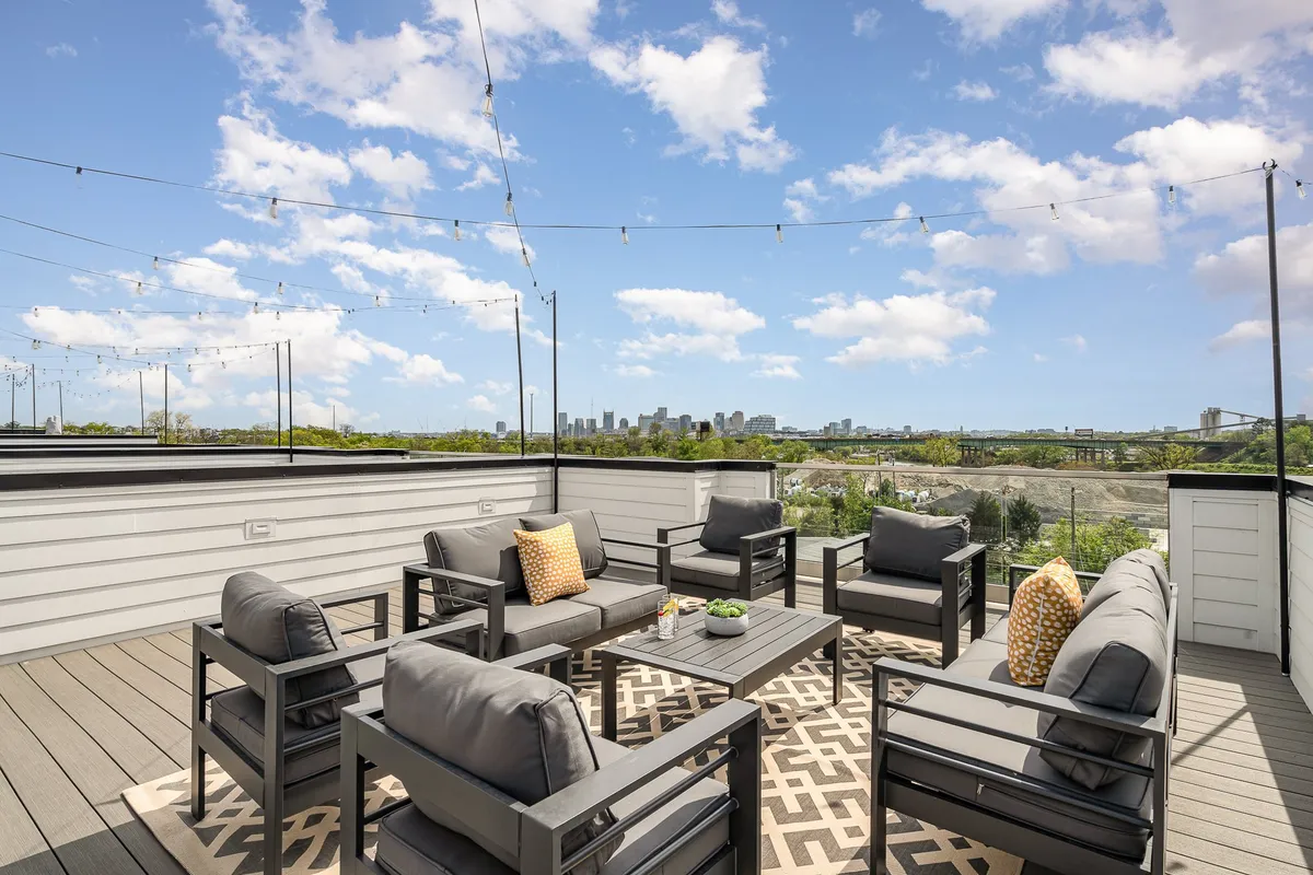 A stylish rooftop seating area overlooks the city skyline. String lights hang above, casting a warm glow over decorative pillows on the cushioned benches. The view stretches across the rooftops to the distant cityscape, inviting both daytime relaxation and evening gatherings.