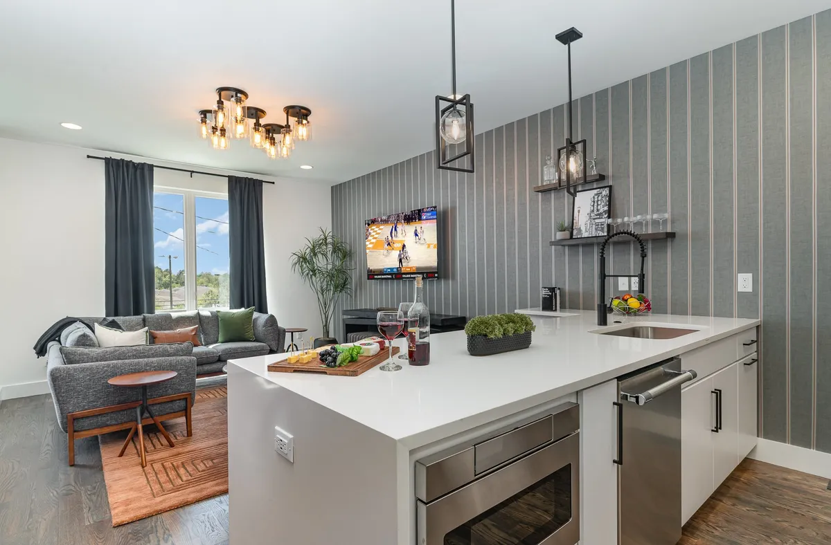 A sleek, contemporary space combining a kitchen island, modern lighting fixture, TV screen, and cozy sofa seating. The layout is perfect for entertaining, with ample room for cooking, mingling, and watching events.